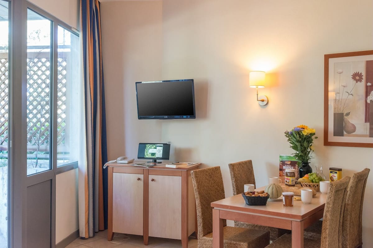 A hotel room with a wooden desk, a flat-screen TV, a wall-mounted lamp, a dining table with chairs, and a window with curtains.