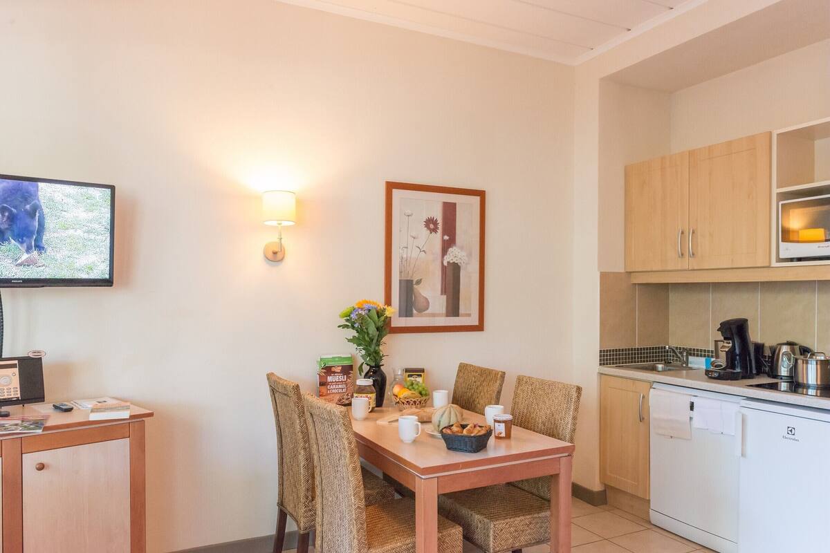 A dining area with a table set for two, a television, a framed picture, and a kitchen with a dishwasher and cabinets.