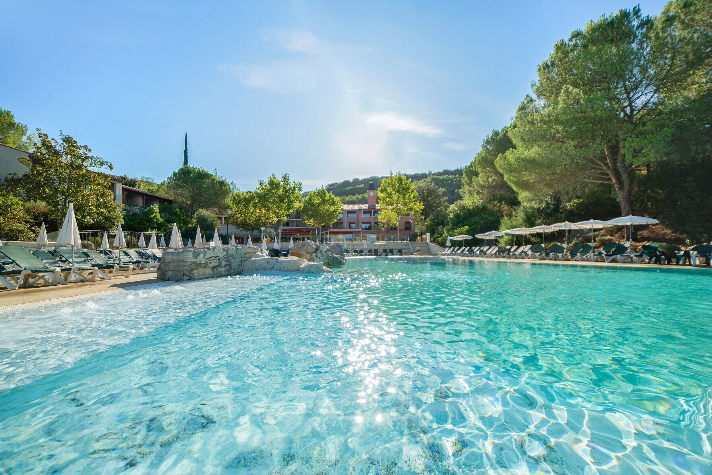 Ein klares, blaues Schwimmbecken mit Liegestühlen und Sonnenschirmen, umgeben von Bäumen und Gebäuden.