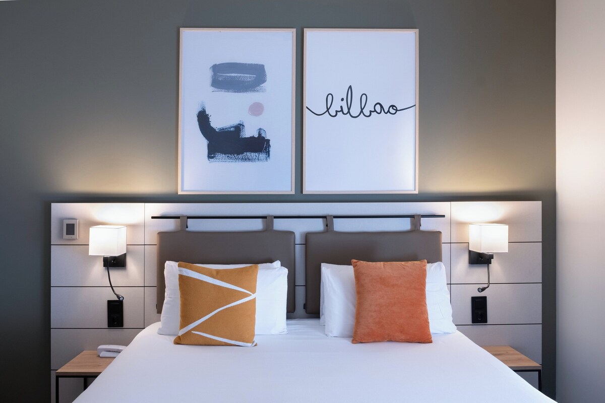 Une chambre d’hôtel avec un grand lit, deux tables de chevet, deux lampes fixées au mur et deux œuvres d’art encadrées au-dessus du lit.