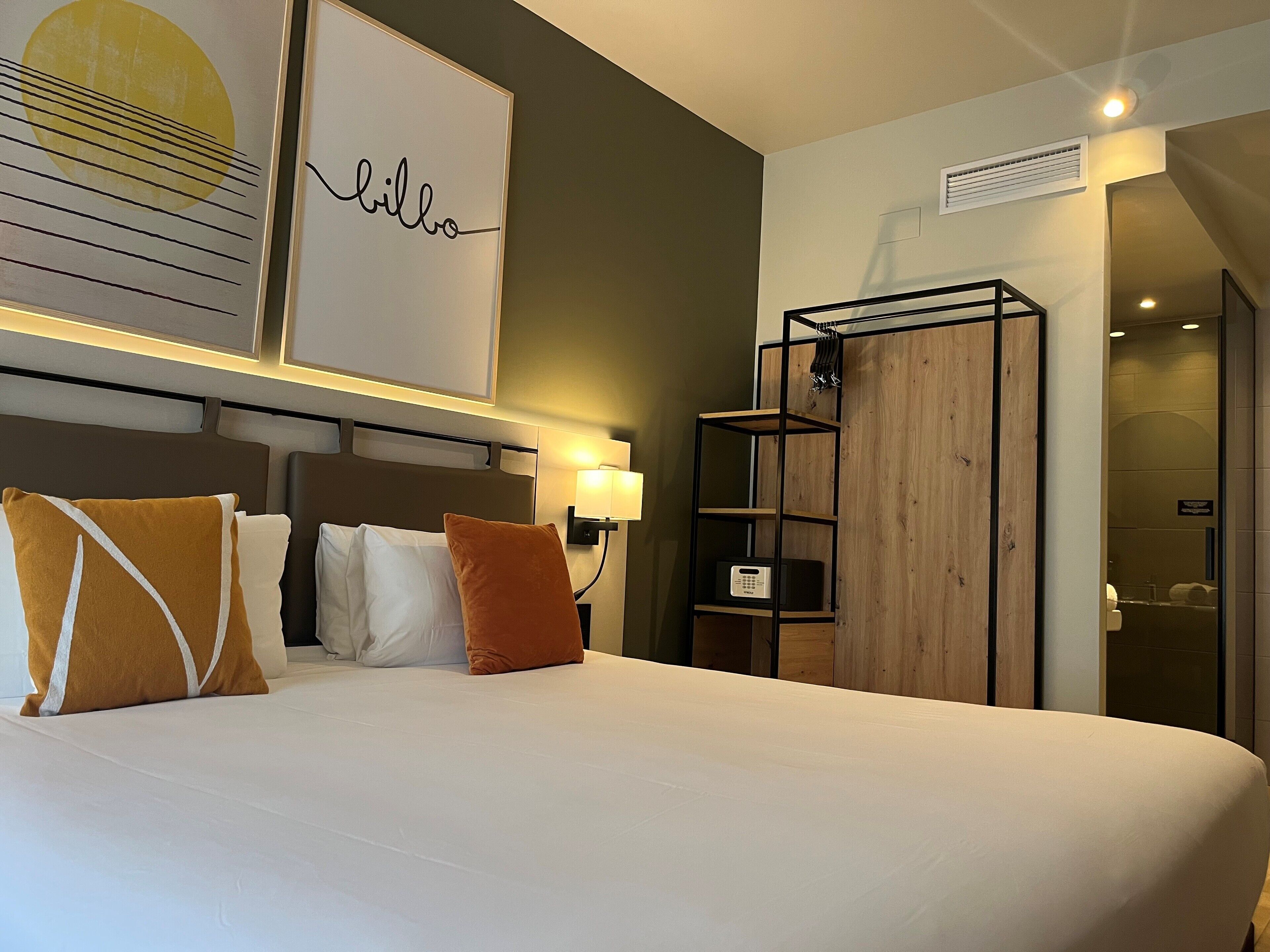 Una habitación de hotel con una cama grande, un cabecero con estante incorporado, una mesita de noche con lámpara y un armario de madera.