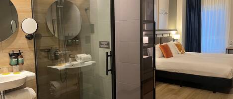 A modern hotel room with a glass-enclosed shower, a bedside table with toiletries, a bed with pillows, and a large window with curtains.