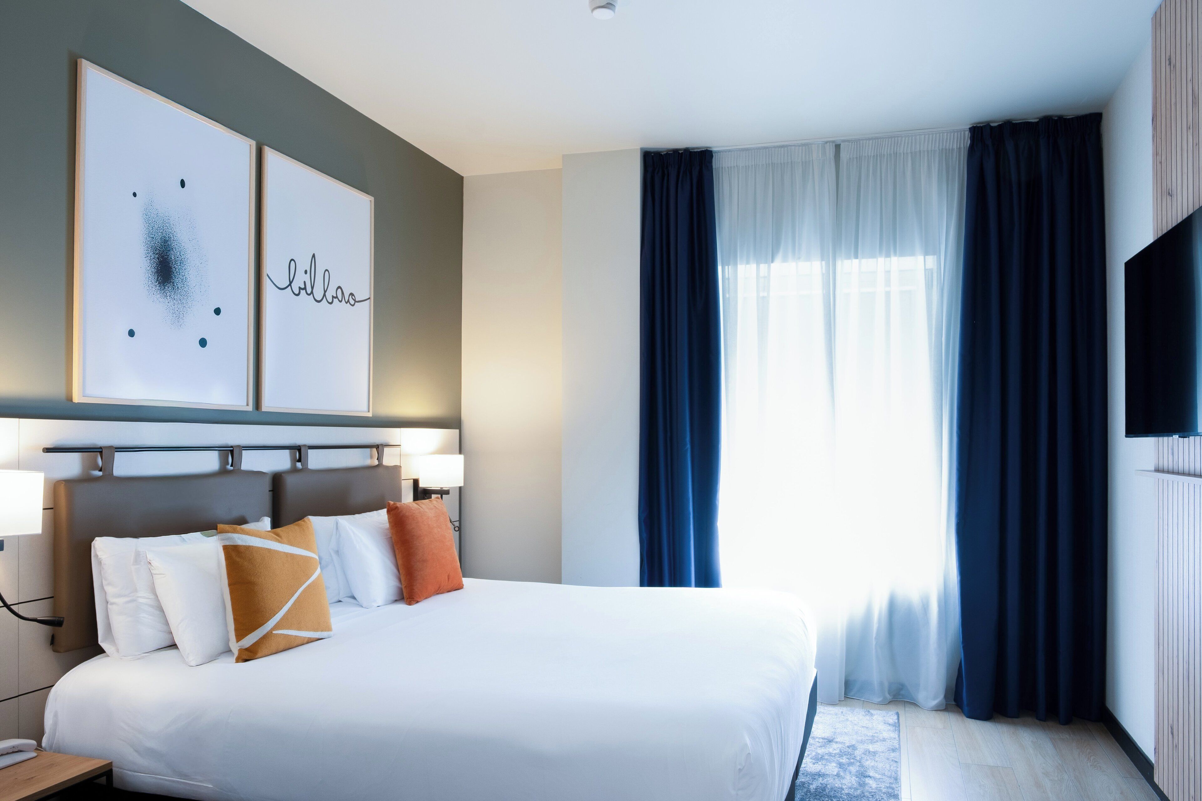 A hotel room with a large bed, two wall-mounted artworks, a flat-screen TV, and a large window with curtains.