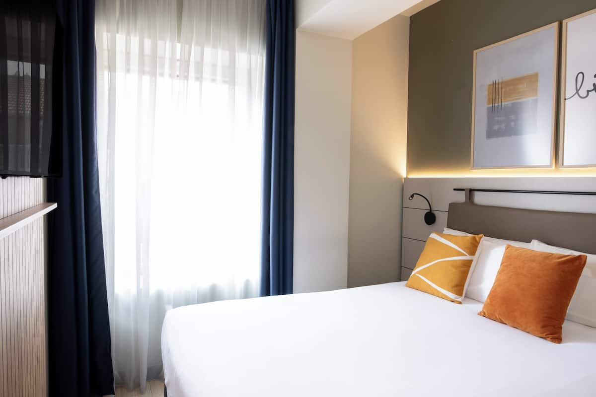 A hotel room with a bed, curtains, a television, and artwork on the wall.