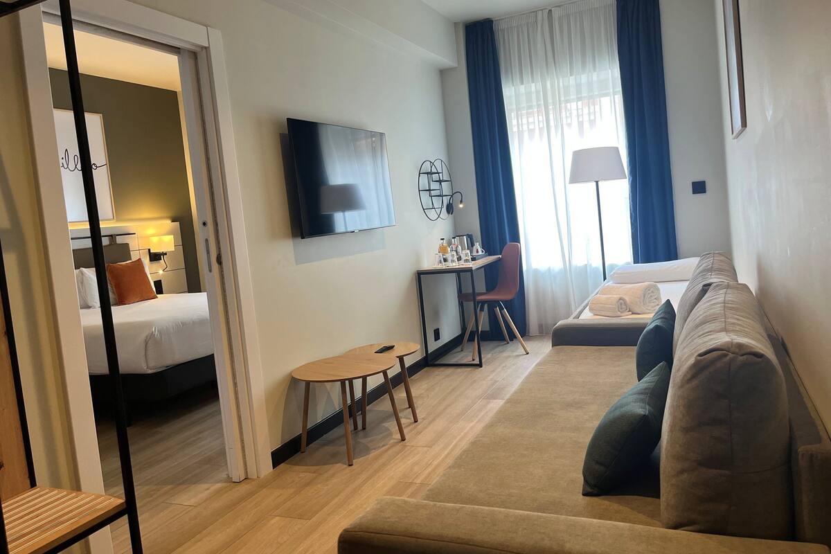 Ein modernes Hotelzimmer mit einem Bett, einem Sofa, einem Schreibtisch mit Stuhl, einem Fernseher und einem Fenster mit Vorhängen.