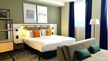 A hotel room with a bed, a sofa, a desk with a lamp, and artwork on the wall.
