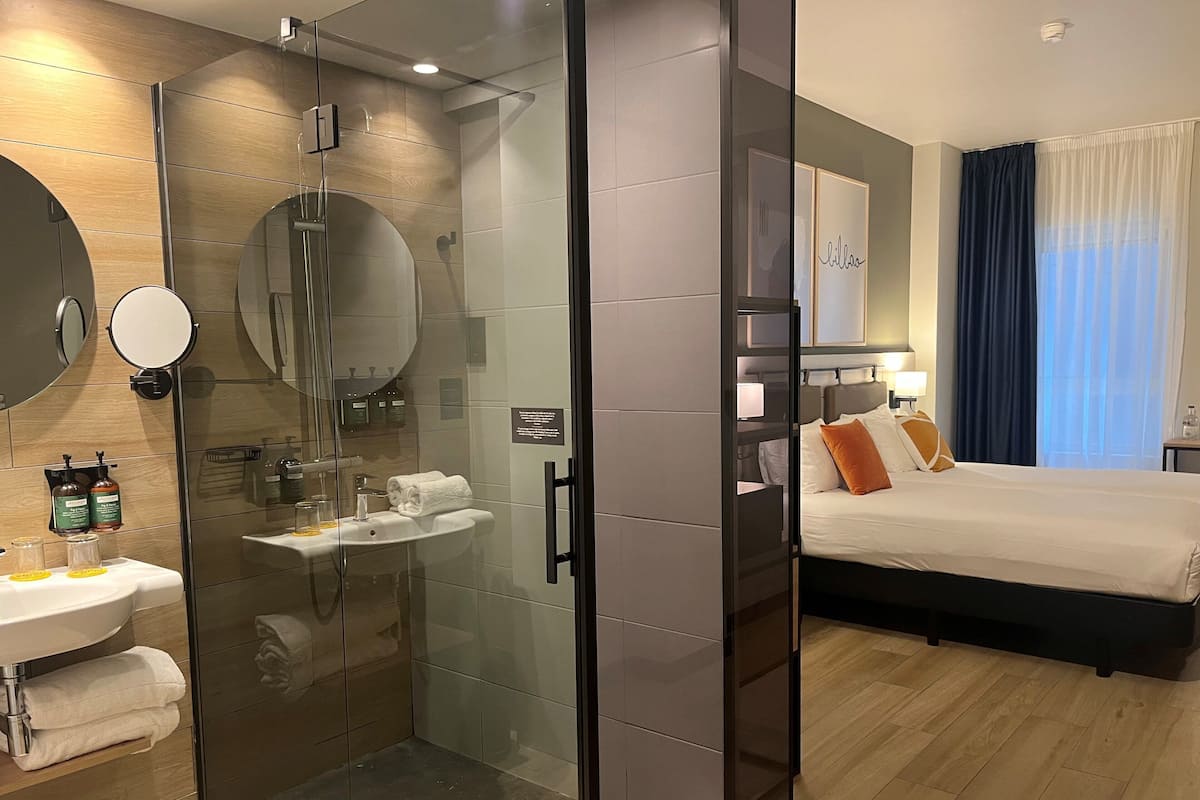 Ein modernes Hotelzimmer mit einer Glaskabine zur Dusche, einem Nachttisch mit Pflegeprodukten, einem Bett mit Kissen und einem großen Fenster mit Vorhängen.