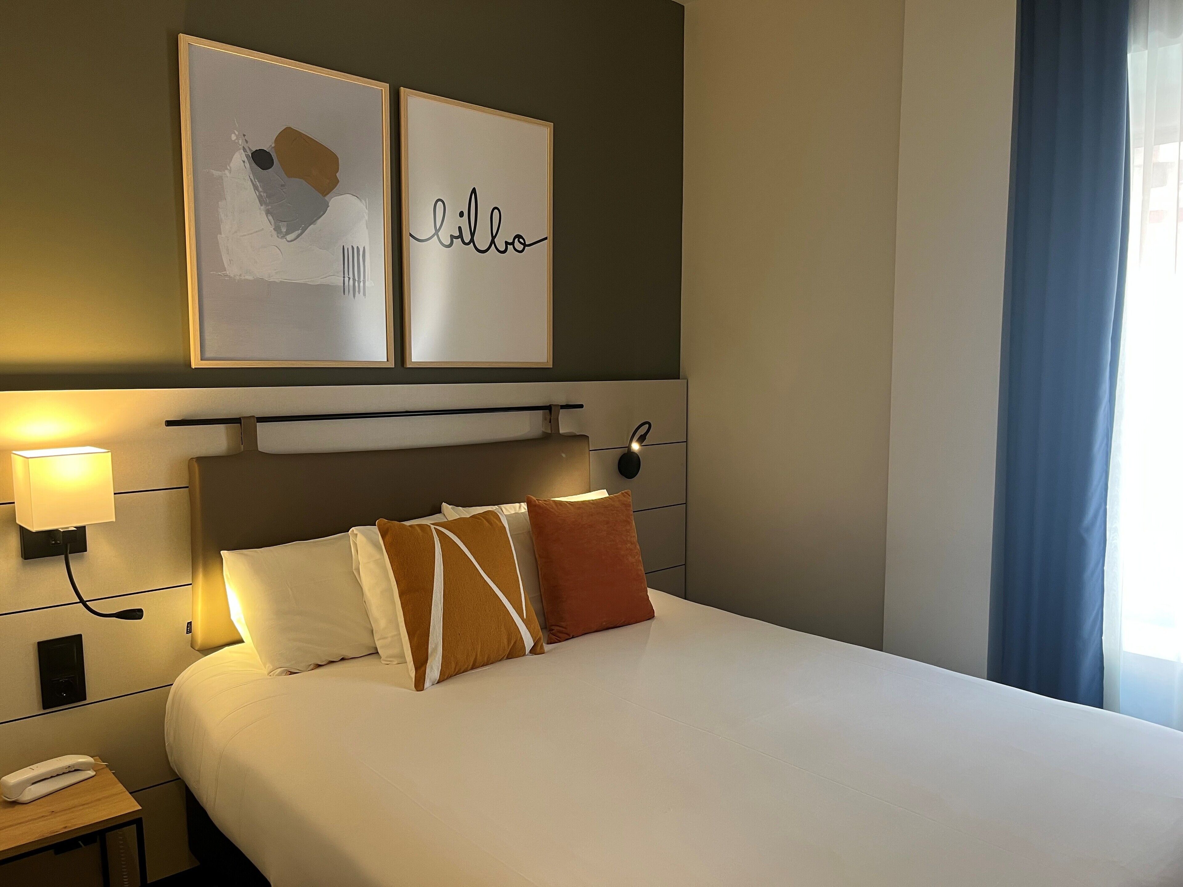 A hotel room with a bed, bedside lamp, phone, and artwork on the wall.