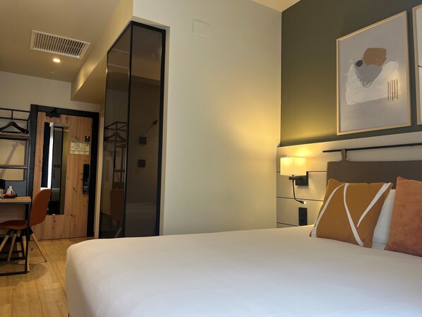 A hotel room with a bed, a desk, a chair, a wardrobe, and a wall-mounted lamp.