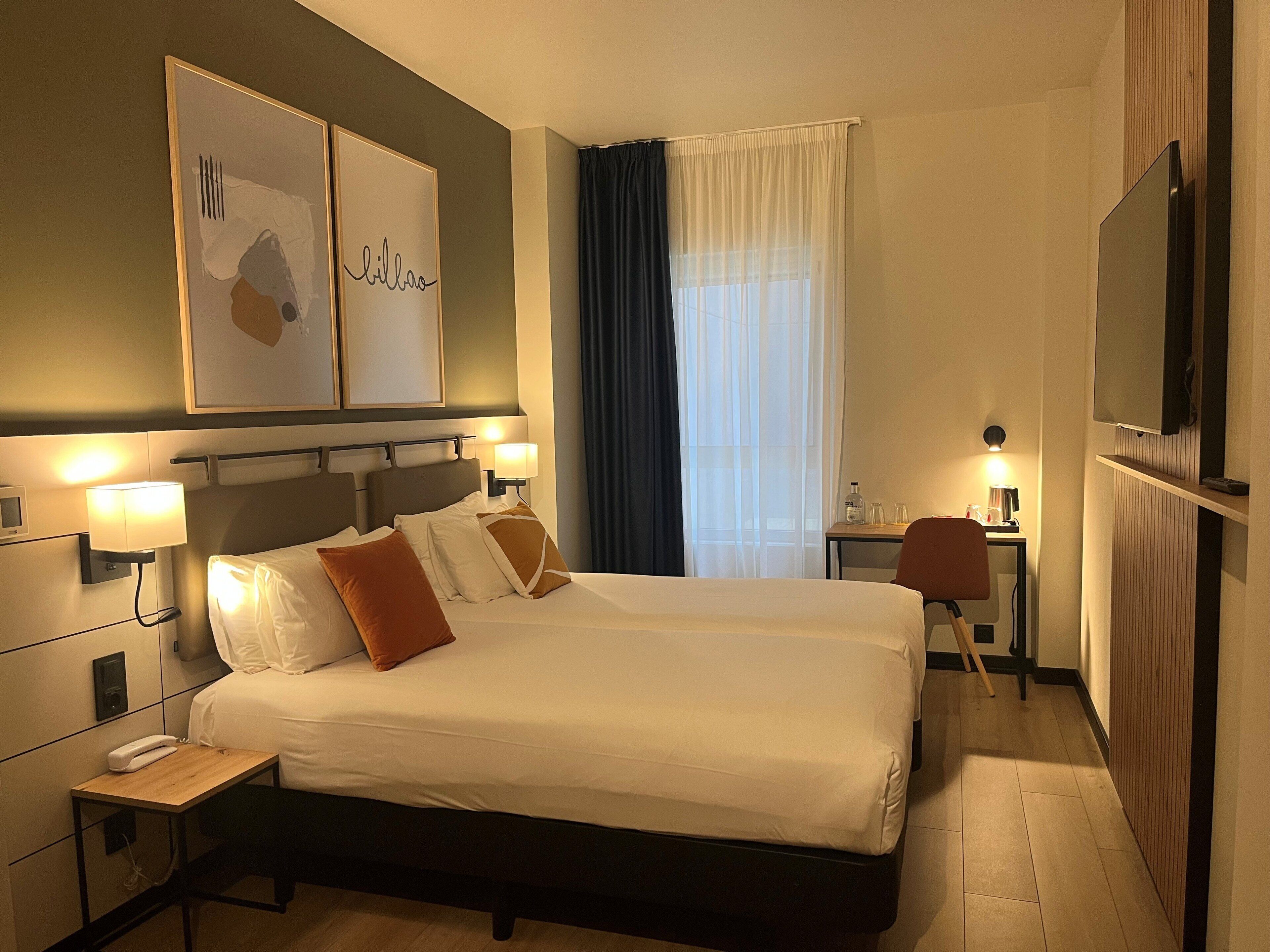 A hotel room with a large bed, bedside lamps, a desk with a chair, a TV, and artwork on the wall.