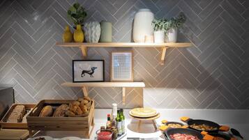 Una cucina con un rivestimento a mosaico a spina di pesce, mensole in legno con oggetti decorativi, un quadro incorniciato e diversi alimenti e utensili da cucina sul piano di lavoro.