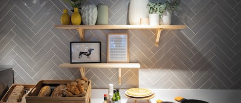 Una cucina con un rivestimento a mosaico a spina di pesce, mensole in legno con oggetti decorativi, un quadro incorniciato e diversi alimenti e utensili da cucina sul piano di lavoro.
