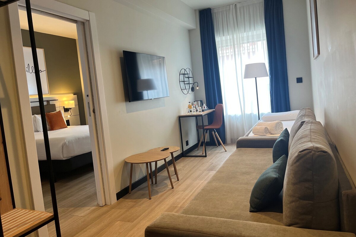 Une chambre d’hôtel moderne équipée d’un lit, d’un canapé, d’un bureau avec une chaise, d’une télévision et d’une fenêtre avec des rideaux.