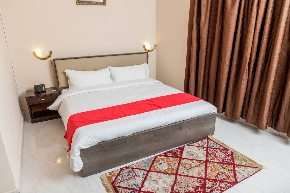 A neatly made bed with a red and white bedspread, a wooden headboard, and a patterned rug on a tiled floor.