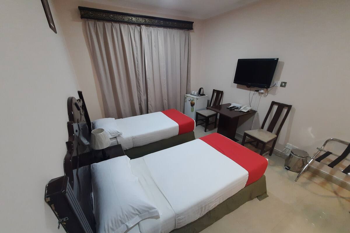 A hotel room with two single beds, a desk with a chair, a television mounted on the wall, and a small table with a kettle.