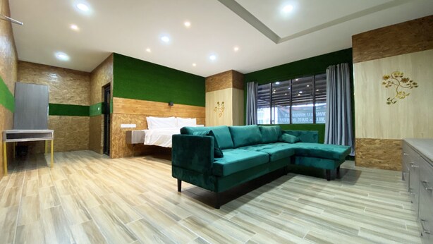 Une chambre d’hôtel moderne avec un canapé d’angle vert, un lit avec du linge de maison blanc, un petit bureau et une grande fenêtre avec des rideaux.