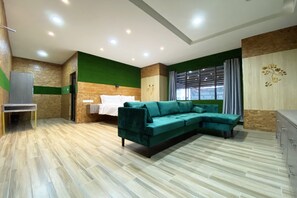 A modern living room with a green sectional sofa, a bed with white linens, a small table, and a large window with curtains.
