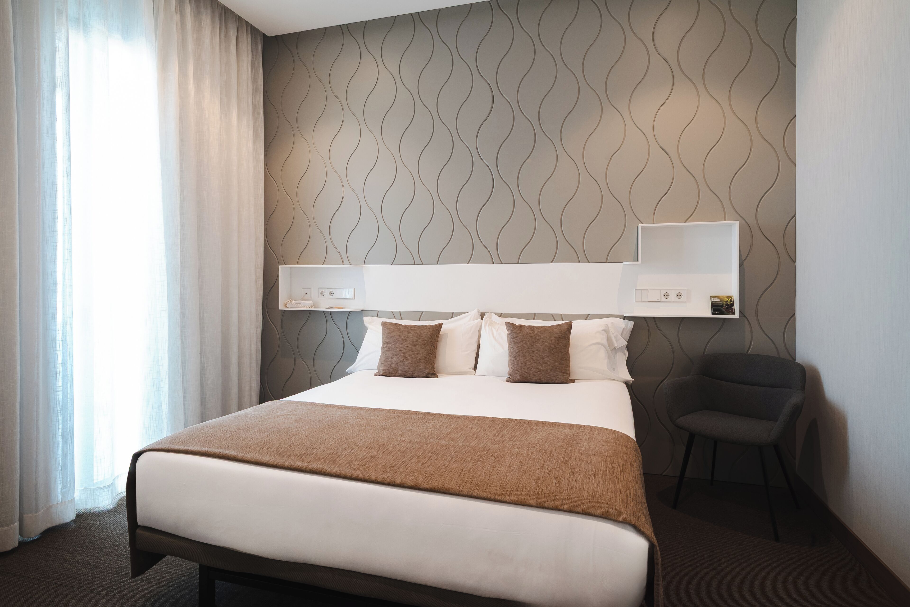 Habitación de hotel con una cama grande, una silla, una mesita de noche y un estante en la pared.
