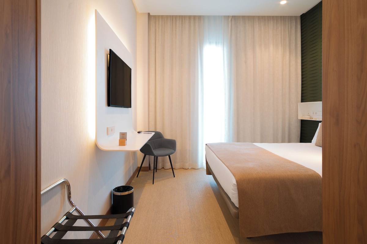 Quarto de hotel com uma cama, uma escrivaninha, uma cadeira e uma televisão na parede.