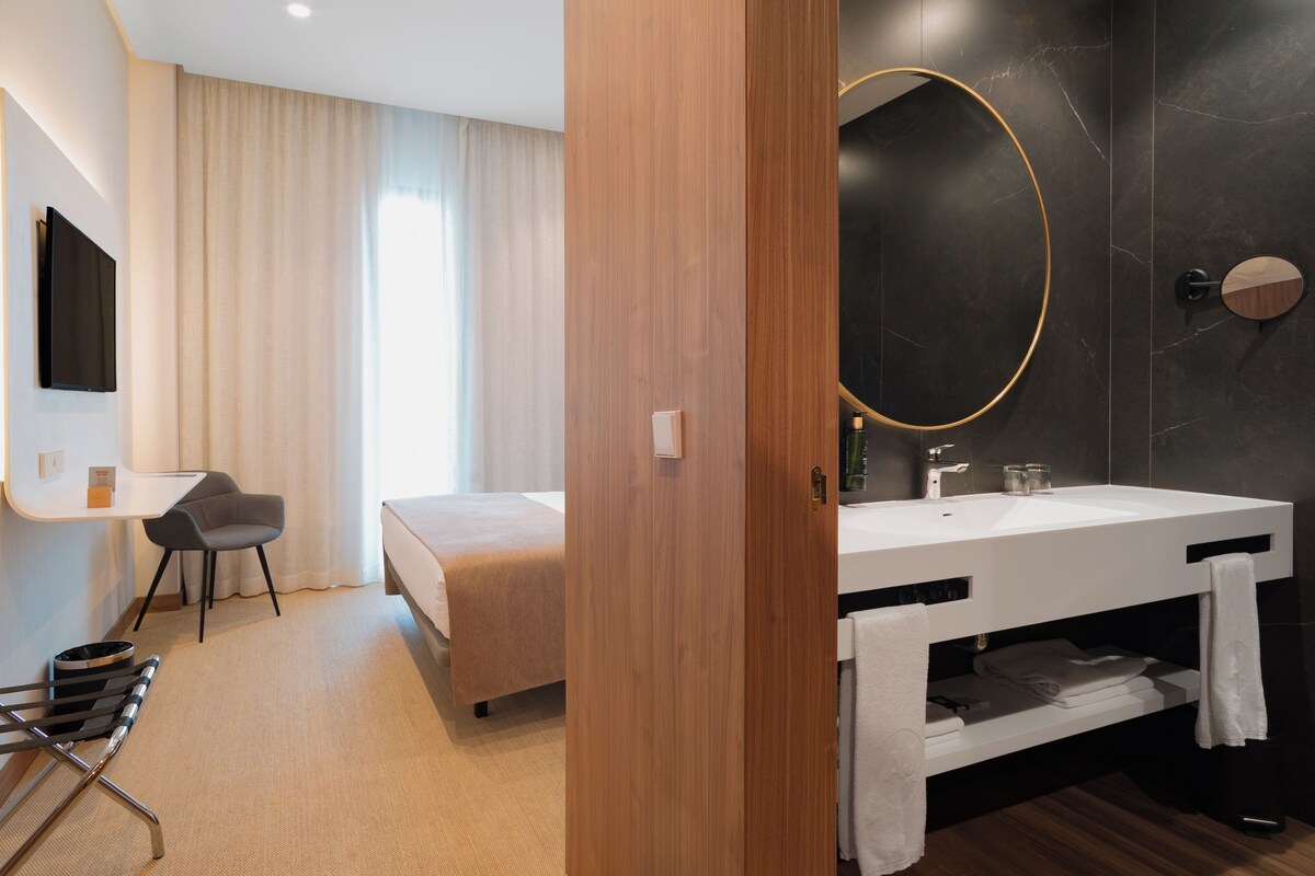 A modern hotel room with a wooden door, a white desk, a grey chair, a bed, a large mirror, and a dark marble wall.