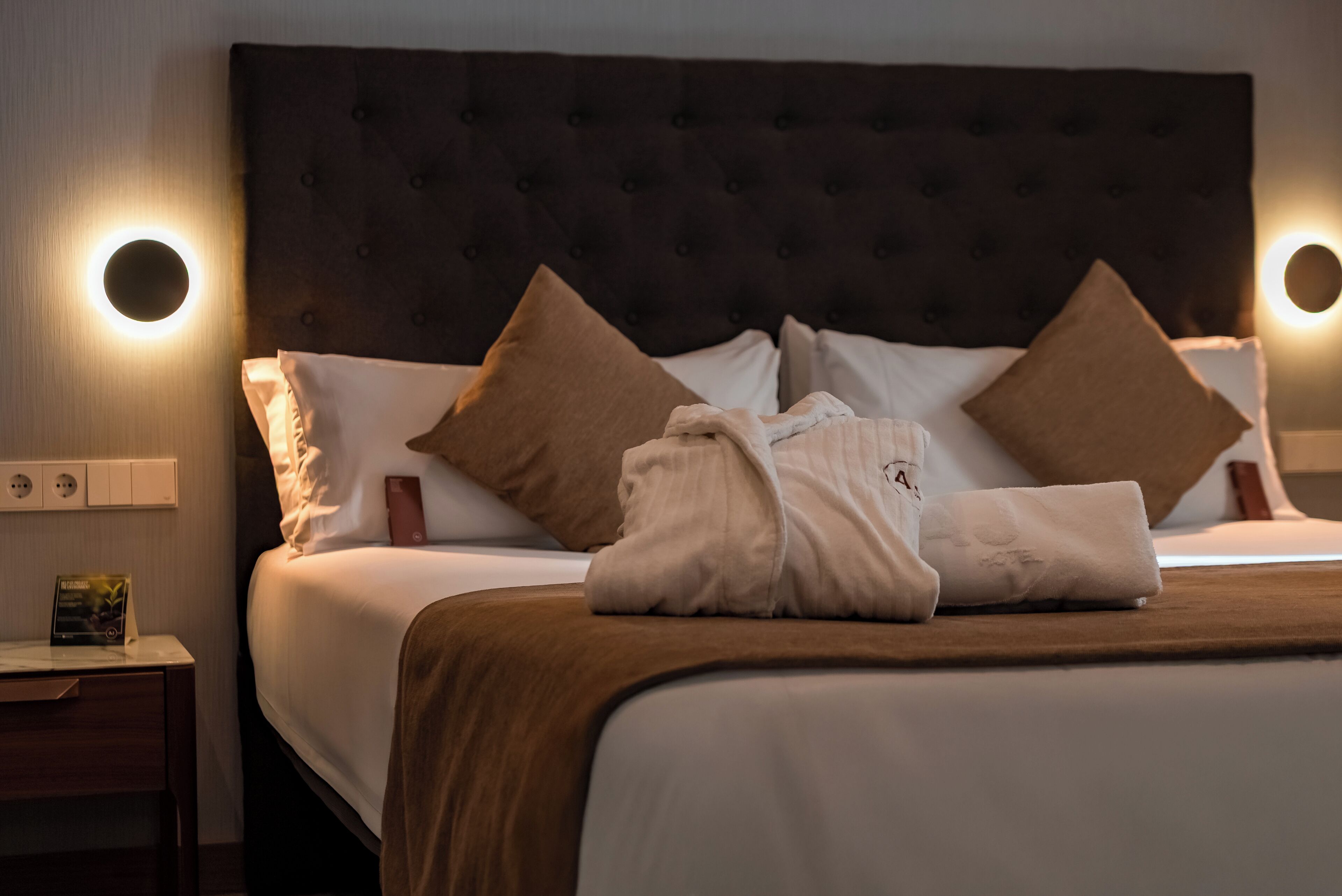 Una habitación de hotel con una cama grande, dos almohadas, una mesita de noche con lámpara y una lámpara de pared.