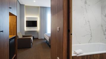 Habitación de hotel con armario de madera, sofá, cama, televisor montado en la pared y baño de mármol con bañera.