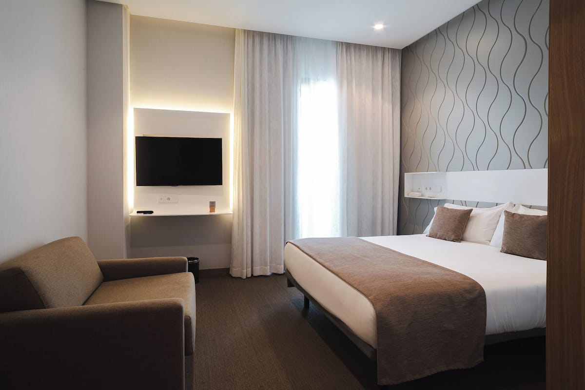 Habitación de hotel con una cama, un televisor montado en la pared, un sofá y una ventana con cortinas.