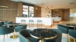 A modern bar area with round tables and bar stools, a visible bar counter, and a variety of bottles in the background. - Hotel AJ Gran Alacant (Santa Pola)