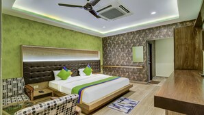 Quarto de hotel moderno com uma cama grande, cabeceira de madeira, parede verde, papel de parede estampado, ventilador de teto, ar condicionado, mesa de cabeceira de madeira, mesa lateral, uma cadeira e uma pequena mesa.