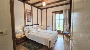 A bedroom with a wooden ceiling, a bed with white bedding, a nightstand with a lamp, a window with curtains, and a door. - Périgord house with fireplace near Monpazier (Mazeyrolles)