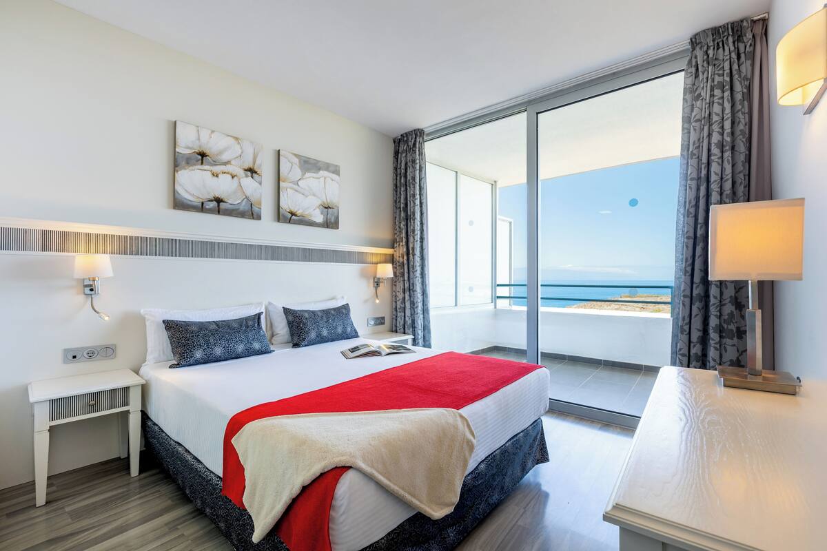 Une chambre d’hôtel moderne avec un grand lit, une table de chevet, une commode et une vue sur la mer par une grande fenêtre.