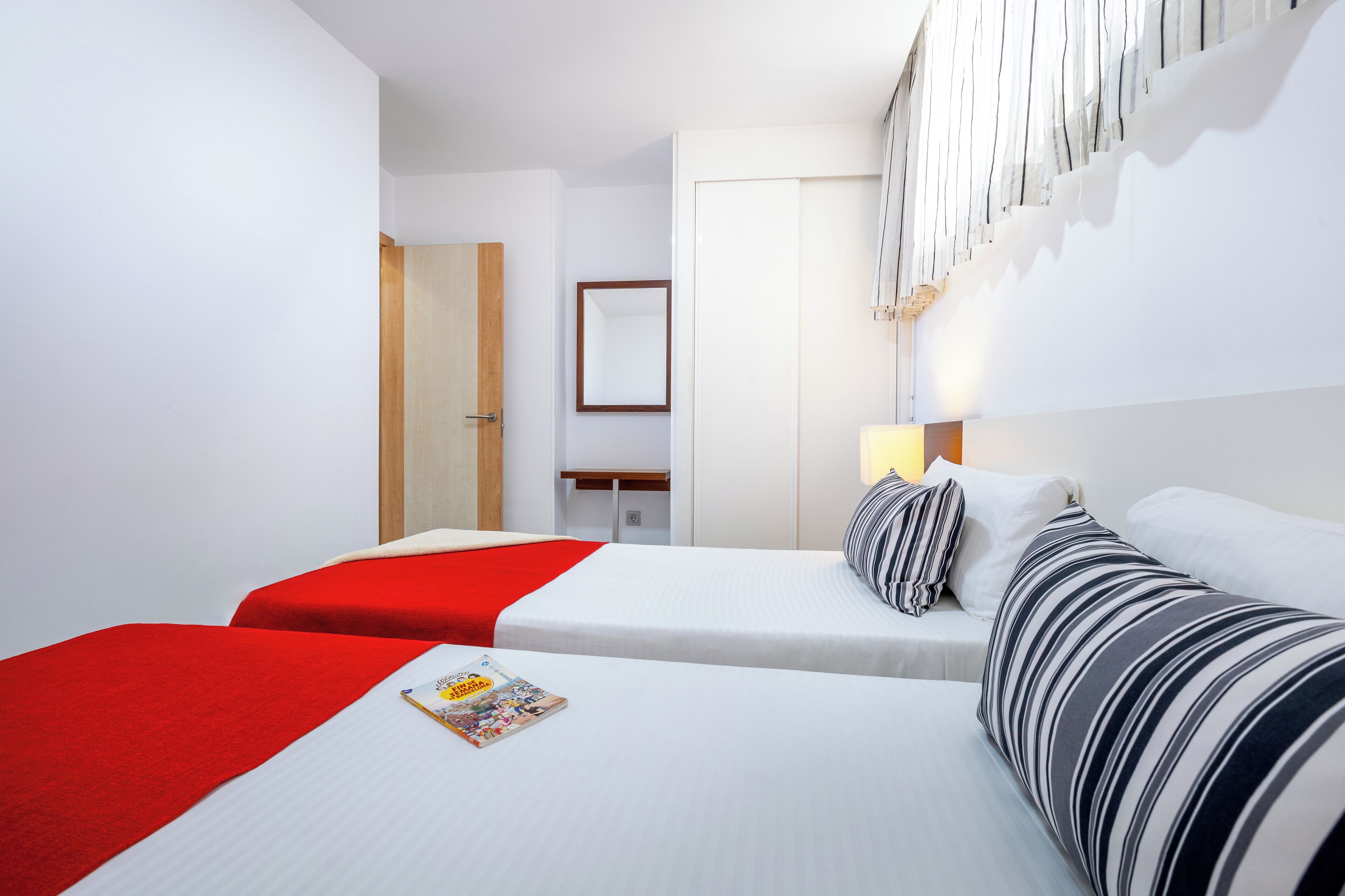 A hotel room with two beds, one with a red bedspread and the other with striped pillows.