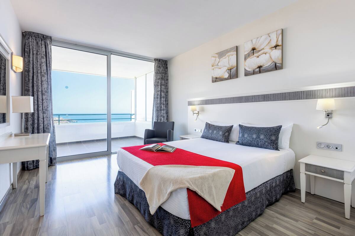 Une chambre d’hôtel moderne avec un grand lit, un bureau et une chaise. De grandes fenêtres offrent une vue sur l’océan.