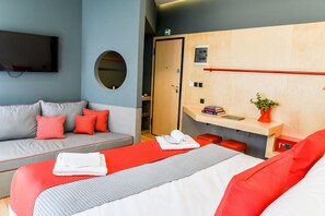 Ett hotellrum med en säng, en soffa, en tv, en spegel, en träpanel med ett rött handtag och en liten växt.