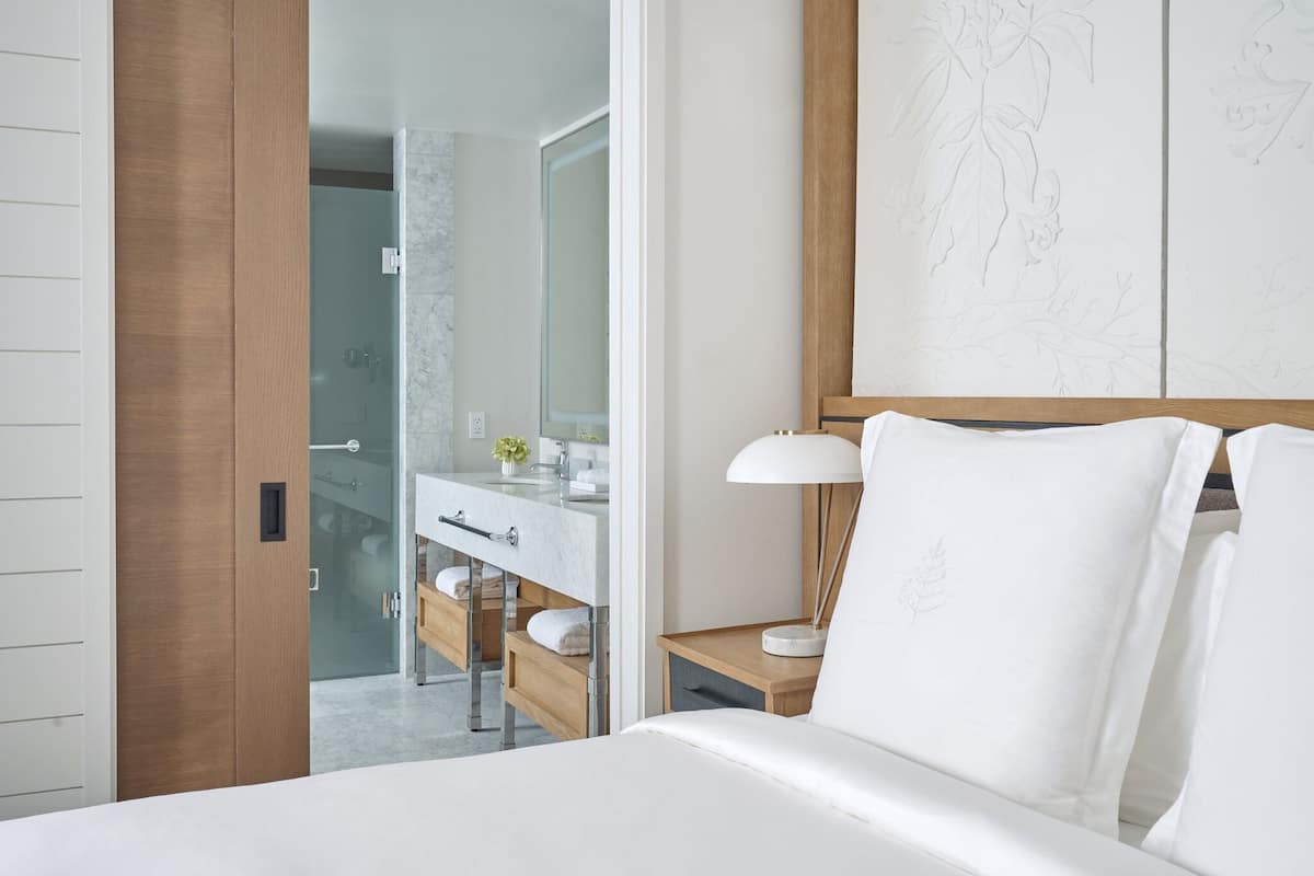 Ein Hotelzimmer mit einem Bett, einem Nachttisch aus Holz, einer weißen Lampe, einem Badezimmer mit Glasdusche und einer Marmorwand.