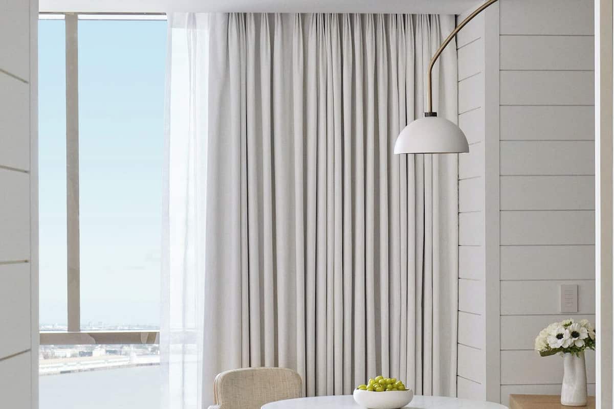 Ein modernes Hotelzimmer mit einem runden Esstisch, zwei Stühlen, einer Lampe und Blick auf die Stadt durch große Fenster.
