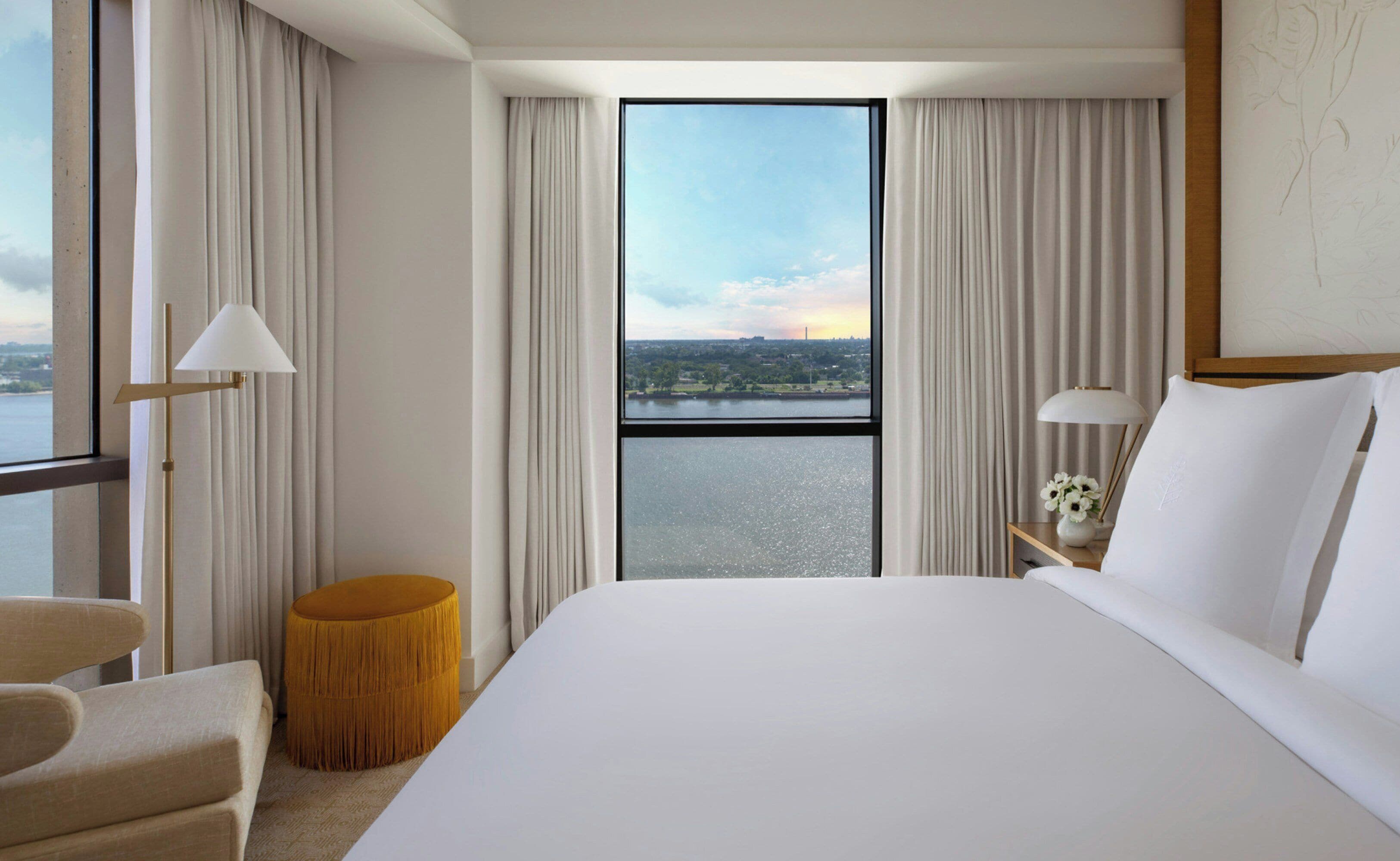 A hotel room with a large bed, a view of a cityscape through a window, and a small round table with a lamp.