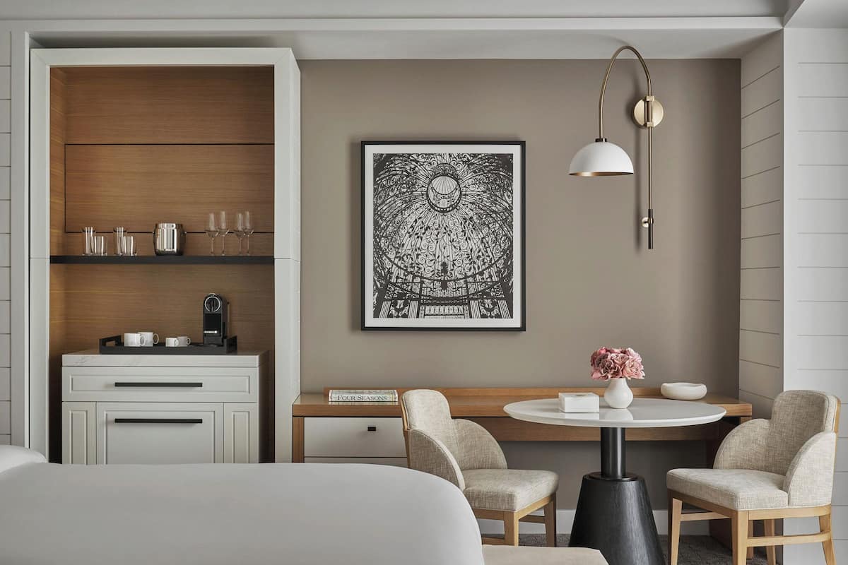 Ein modernes Hotelzimmer mit einem Holzkabinett, einem gerahmten Kunstwerk, einem runden Tisch mit Stühlen und einer Kaffeemaschine.