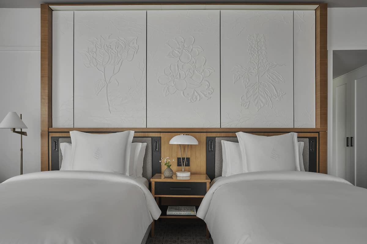 Ein Hotelzimmer mit zwei Betten, einem hölzernen Kopfende mit floralen Verzierungen, einem Nachttisch mit Lampe und einer kleinen Vase mit Blumen.