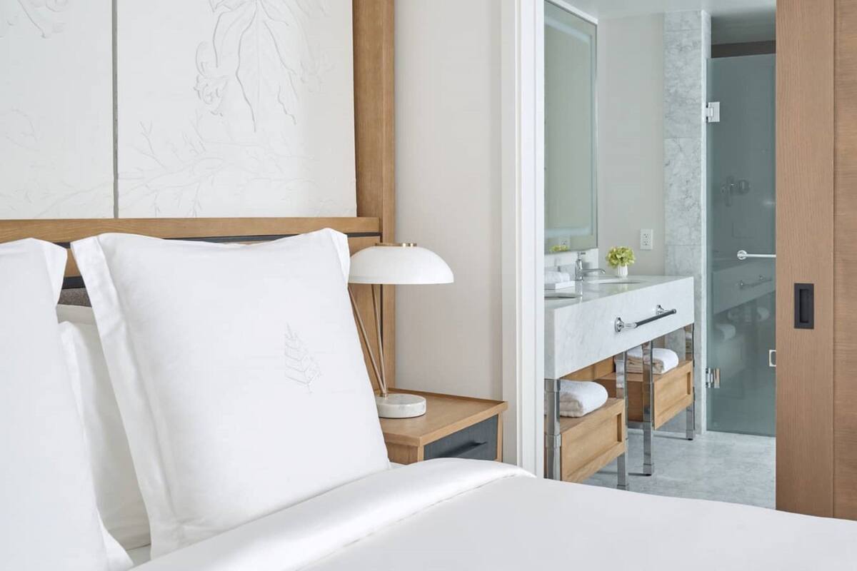 Ein Hotelzimmer mit Bett, Nachttischlampe, Holz-Nachttisch, Badezimmer mit Glasdusche und Holztür.