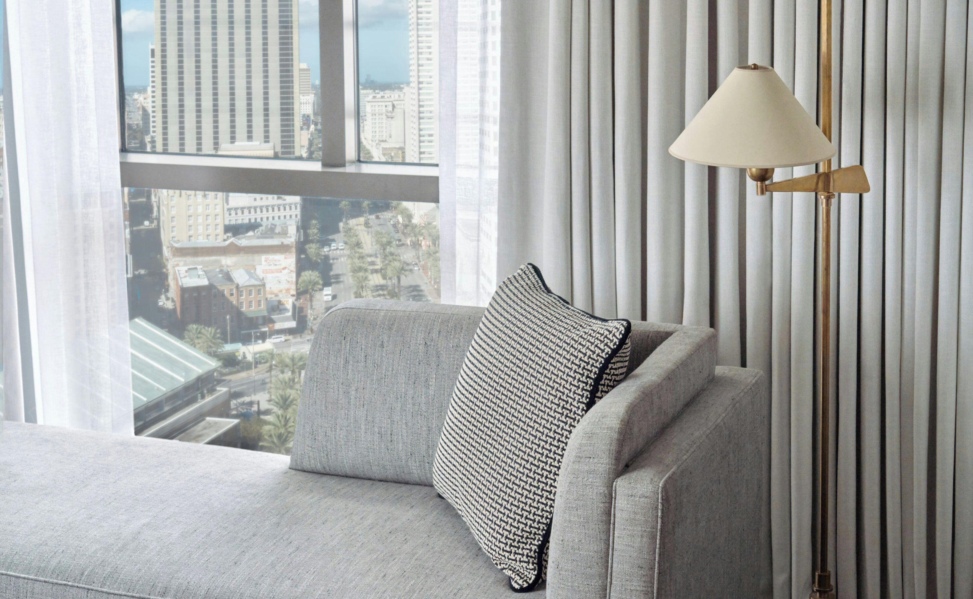 Una sala de estar moderna con un sofá, un cojín estampado, una lámpara de pie y vistas de un paisaje urbano a través de amplios ventanales.
