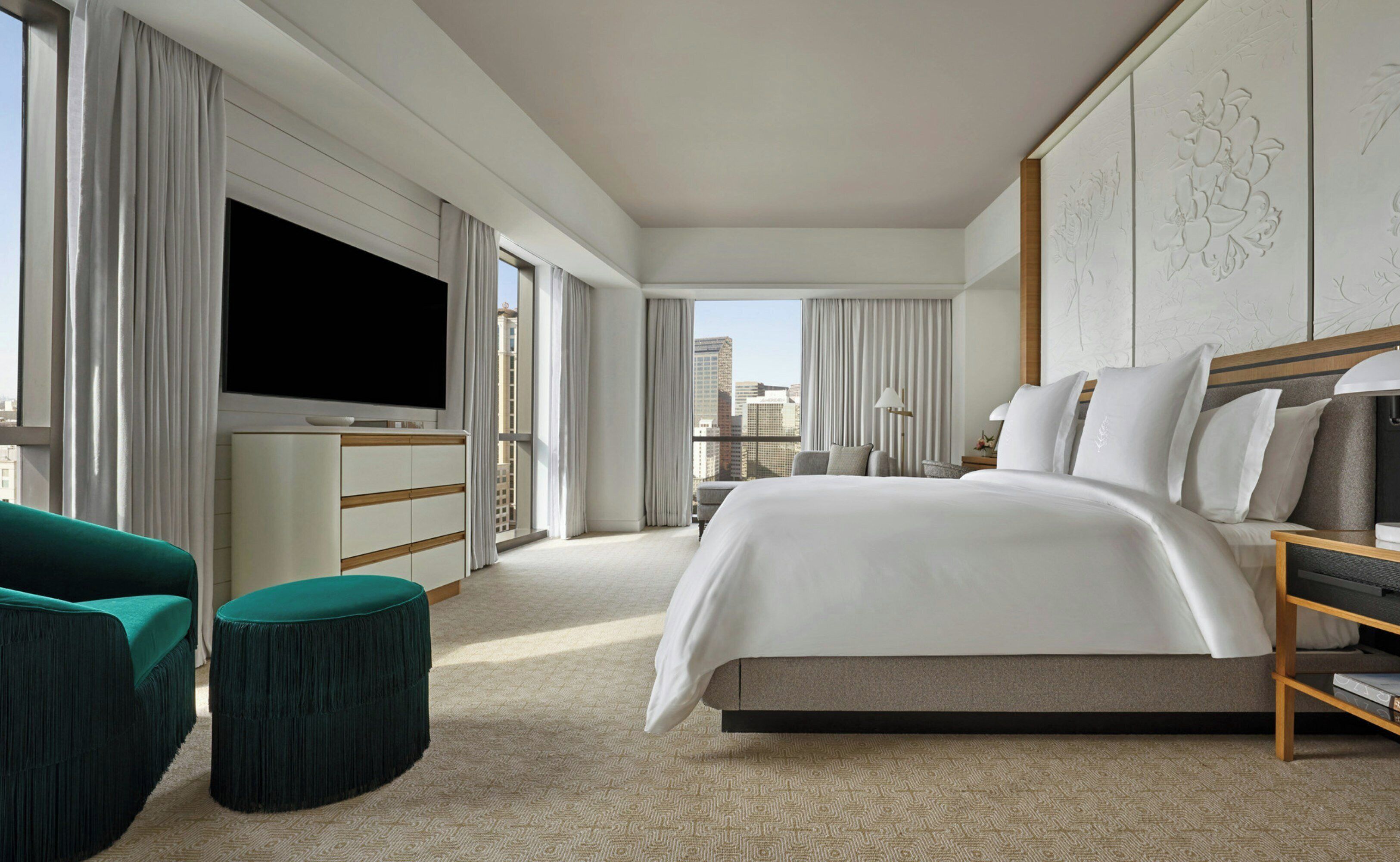 Quarto de hotel moderno com uma cama grande, televisão de tela plana, área de estar com uma poltrona verde e vista para a cidade pelas janelas.