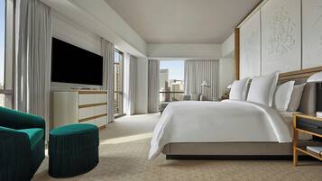 Quarto de hotel moderno com uma cama grande, televisão de tela plana, área de estar com uma poltrona verde e vista para a cidade pelas janelas.