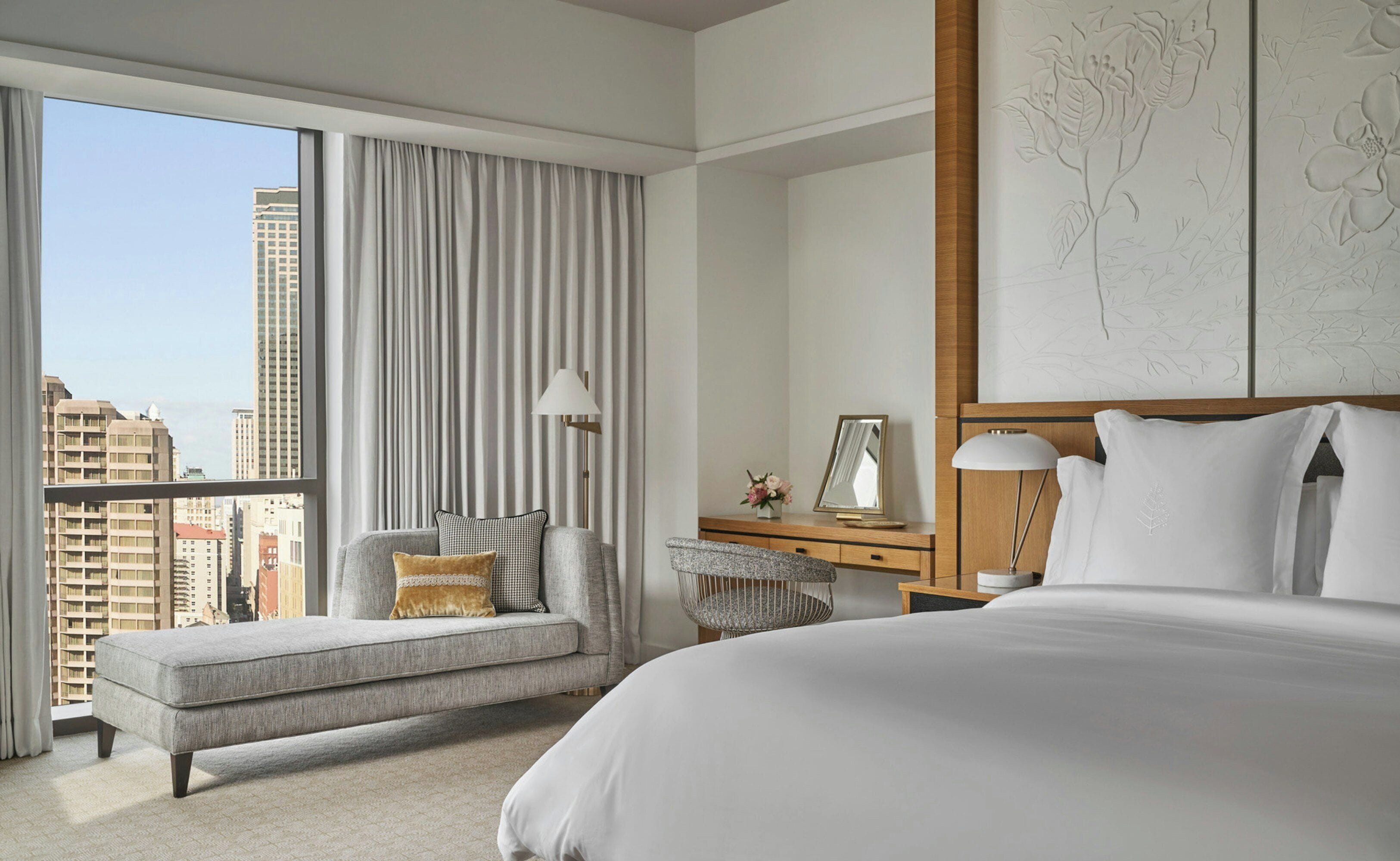 A hotel room with a large bed, a seating area with a view of the city, a desk with a lamp, and a wall with floral artwork.