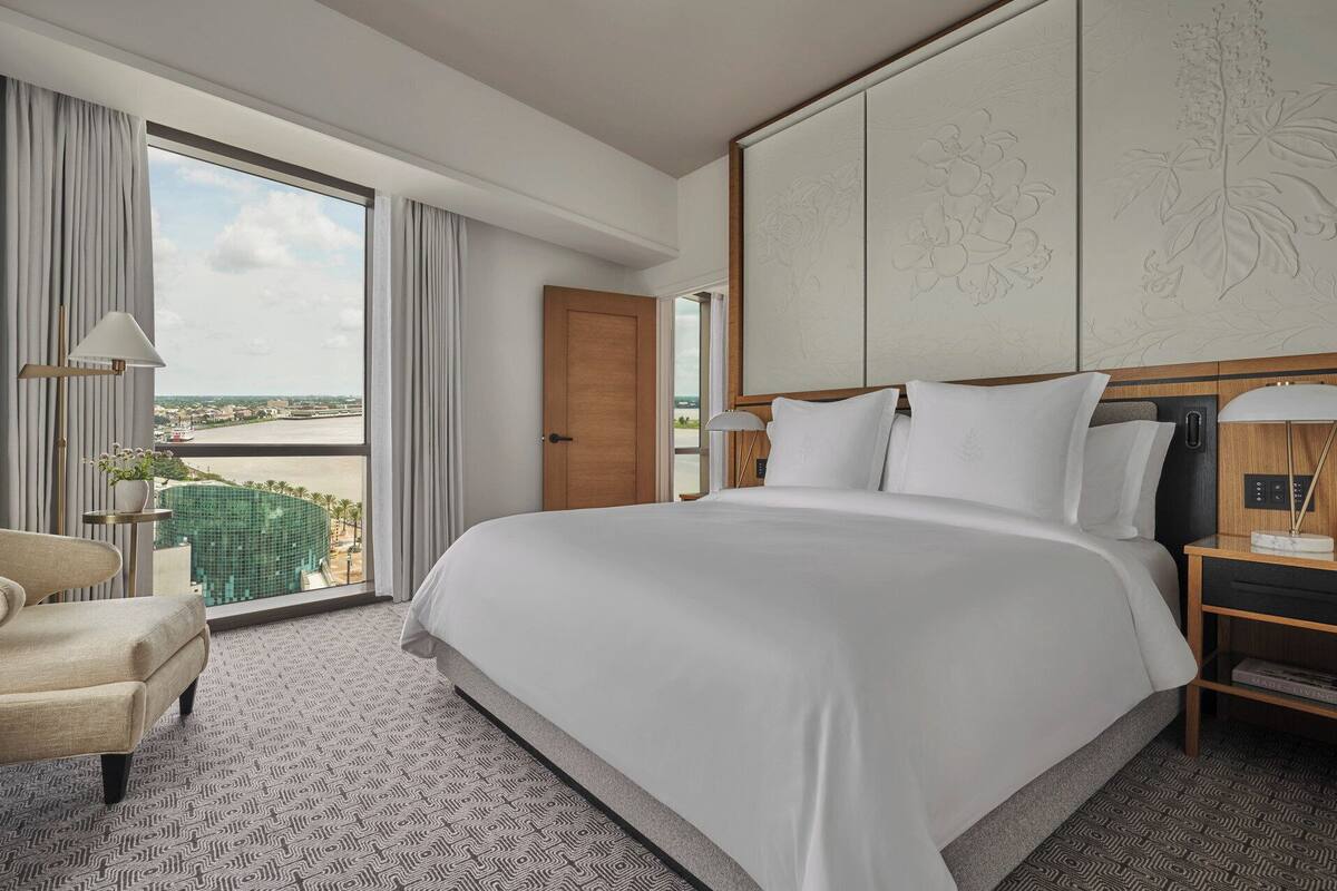 Ein Hotelzimmer mit einem großen Bett, einem Sessel, einem Schreibtisch mit Lampe und Blick auf die Stadt durch große Fenster.