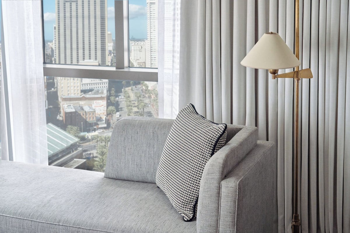 Ein modernes Wohnzimmer mit einer Couch, einem gemusterten Zierkissen, einer Stehlampe und Blick auf eine Stadtlandschaft durch große Fenster.