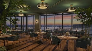 A restaurant with large windows offering a city view, wooden flooring, and modern chandeliers.