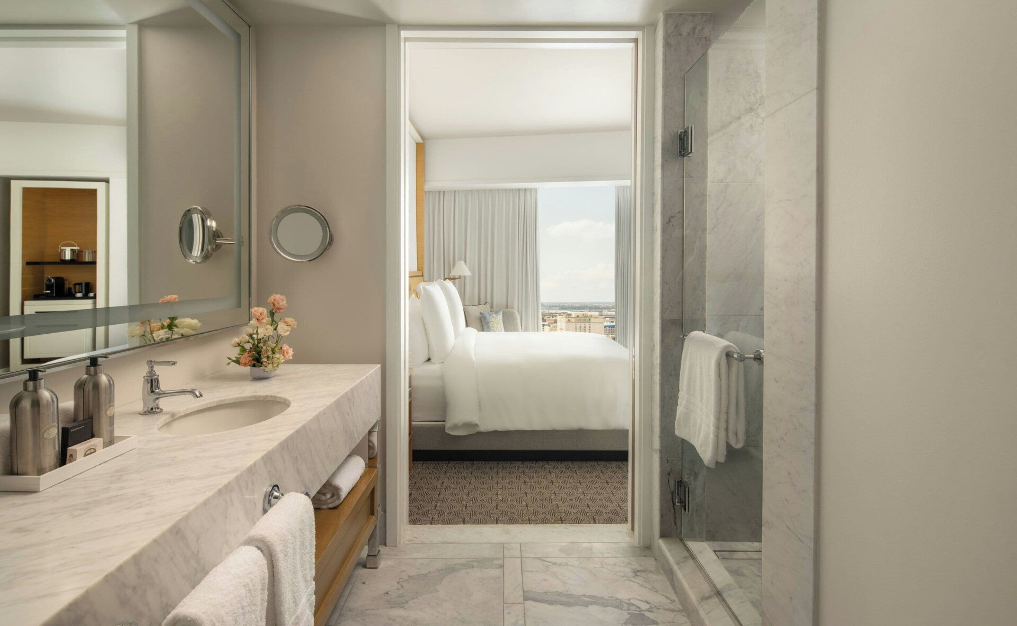 A hotel bathroom with a marble sink, a large mirror, a shower area, and a view of the city through the window.