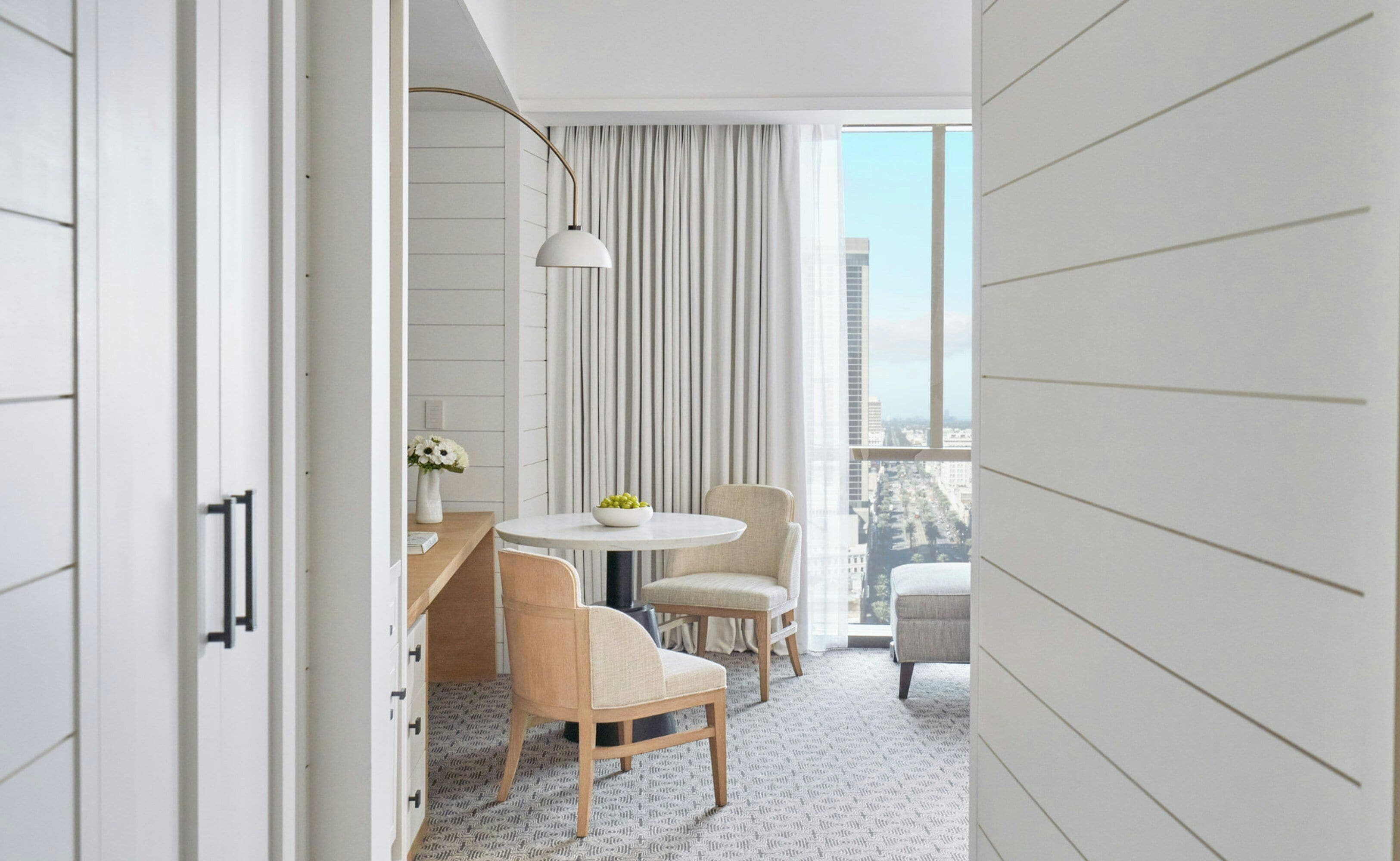 Habitación de hotel moderna con balcón, una mesa redonda, dos sillas, un florero con flores y vistas a la ciudad.