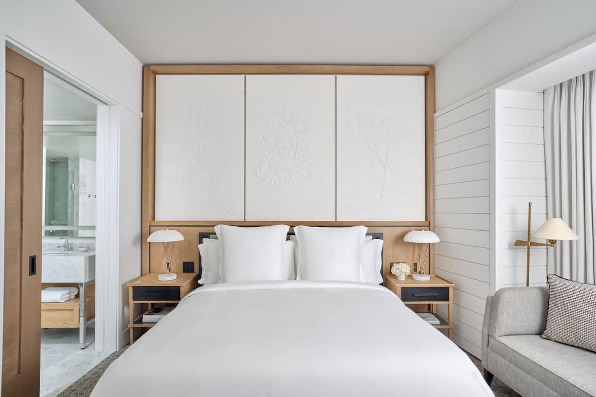 Ein modernes Hotelzimmer mit einem großen Bett, einem Holzkopfende, zwei Nachttischen mit Lampen, einem grauen Sofa und einer großen Wandverkleidung mit floralen Mustern.